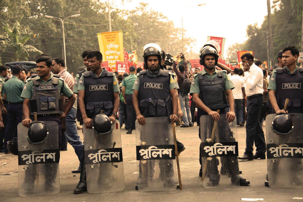 Riot police with shields and helmets standing behind a protesting crowd. 