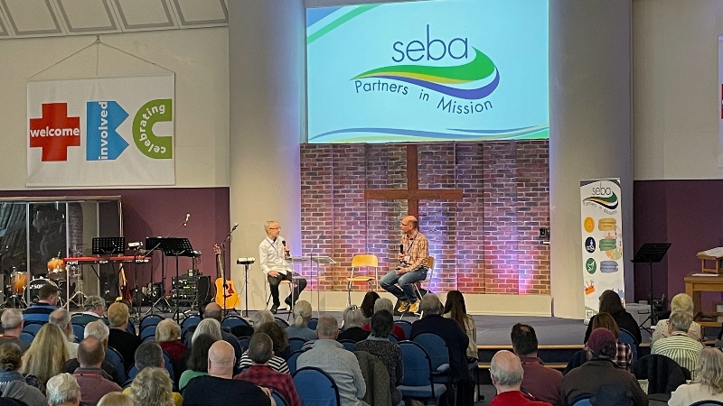 Regional ministers Andy Twilley and Joth Hunt talking on stage watched by conference delegates below a large SEBA Partners in Mission image 