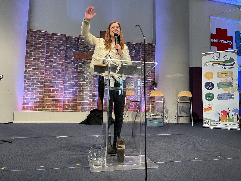 Anne Calver speaking at a lecturn with a microphone in her left hand, and right arm raised