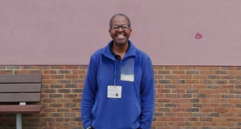 Wayne Dixon stand smiling, hands in pockets, in front of a school wall and bench