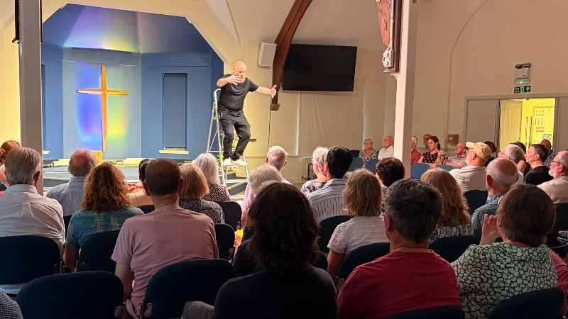 Matt Britton performs the Gospel of Mark on a stage, stood on a step ladder, watched by dozens of people