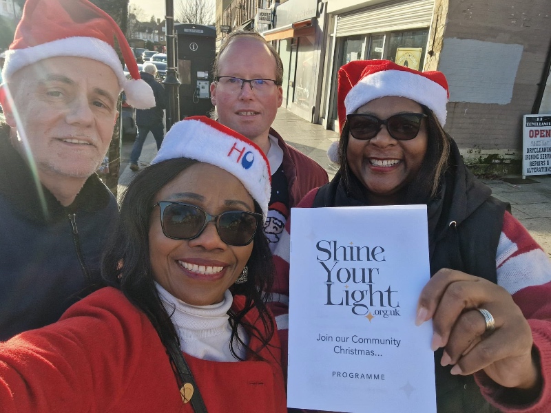 4 people (2 men and 2 women) wearing Santa hats and holding a programme saying 'Shine Your Light' take a selfie on the high street