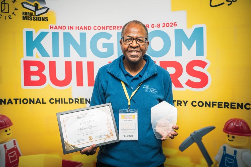 A smiling Wayne Dixon holds his awards - one a certificate, one a glass engraving, in front of a yellow conference banner containing the words "Kingdom Builders"