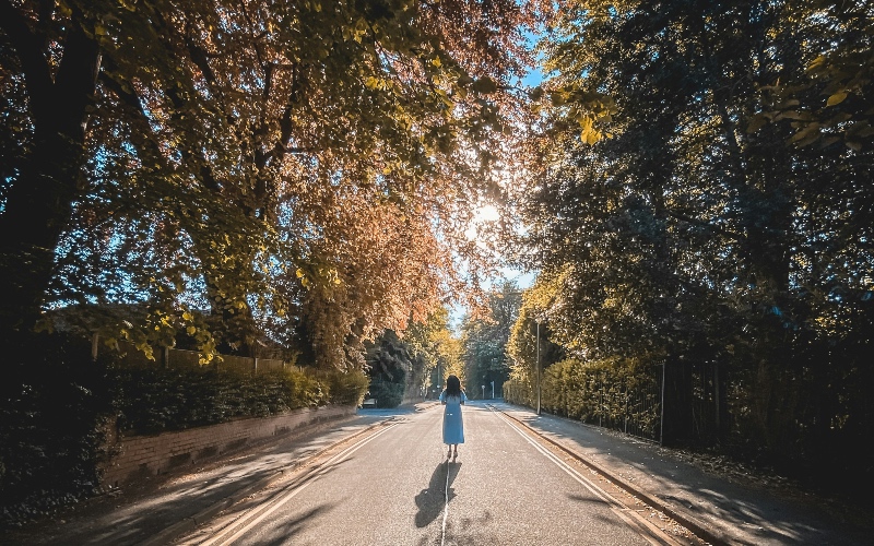 A woman in a white dress stands in the middle of a sun lit, tree lined road 
