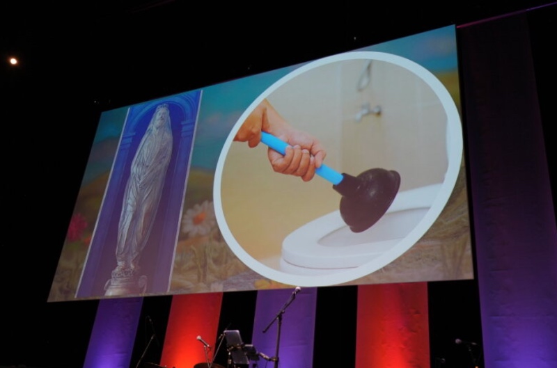 A large screen displays a surreal image of a hand using a plunger in a toilet, beside a statue. Below, stage lights cast purple and red hues.