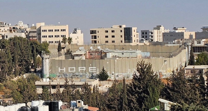 A concrete barrier with "Hope" painted on it stretches across an urban area, surrounded by trees and buildings under a clear blue sky