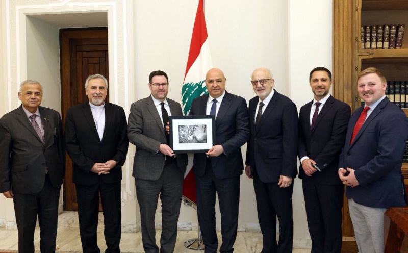 A group of seven men in formal suits standing in a line indoors, with two men in the center holding a framed picture together in front of the flag of Lebanon.
