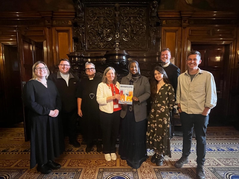 A group of seven people standing together in a richly paneled wooden hall, with two women in the center holding up a colourful booklet titled "MANIFESTO."