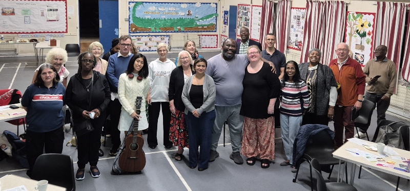 A diverse group of people posing together inside a community hall, with one person holding an acoustic guitar