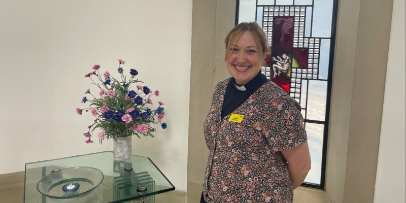 Chaplain Amanda Pink smiling in a chapel room beside a flower arrangement and a stained-glass window